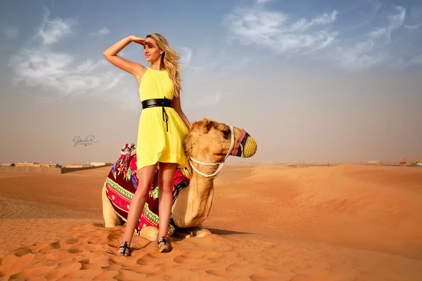 Guest in a yellow dress beside a resting camel on the red dunes